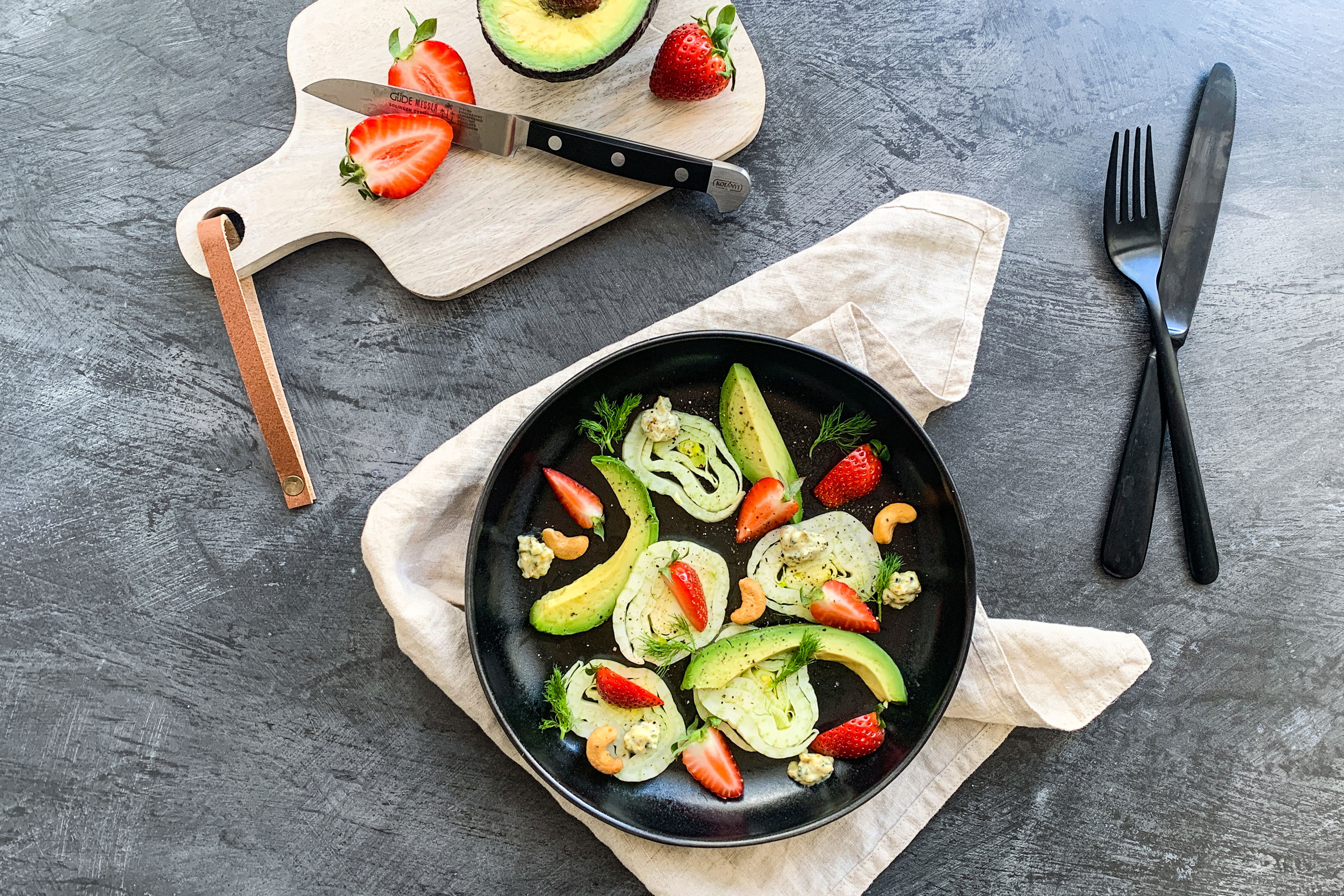 Avocado-Fenchel-Salat mit Kokosdressing in einem tiefen, schwarzen Teller neben einem Brettchen mit Erdbeeren und Avocado